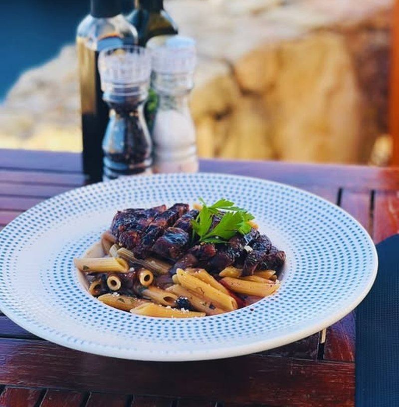 Penne in a dark slow ragù on a timber table by the sea